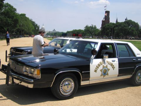 1991 Ford LTD Crown Victoria - man stands beside a black-and-white Ford Highway Patrol sedan parked on the National Mall with the Capitol in the distance.