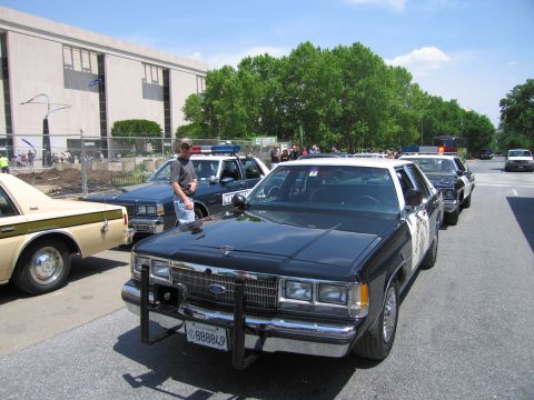 1991 Ford LTD Crown Victoria - black-and-white Ford police sedan with a chrome push bar sits at the front of a lineup of vintage patrol cars at an outdoor show.