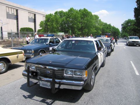 1991 Ford LTD Crown Victoria - black-and-white Ford Highway Patrol sedan with a push bar faces the camera among a row of vintage police cars on a paved lot.