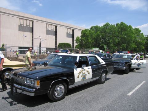 1991 Ford LTD Crown Victoria - black-and-white Highway Patrol Ford sedan leads a line of vintage patrol cars parked along a paved lot beside a large building.