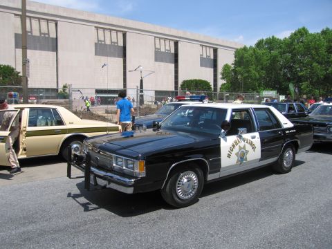 1991 Ford LTD Crown Victoria - black-and-white Ford Highway Patrol sedan with a push bar is parked beside a tan sedan in front of a large stone museum building.