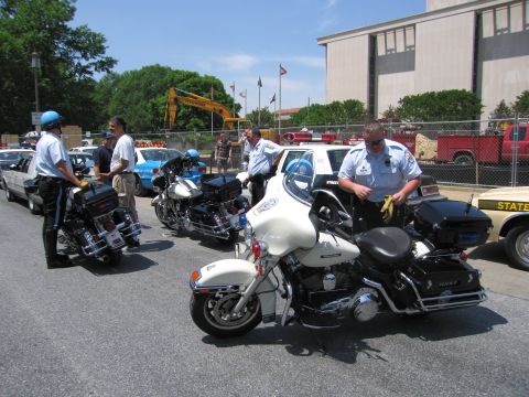 1991 Ford LTD Crown Victoria - Police officers in white short-sleeve uniforms stand among parked white police motorcycles in a paved lot near a building.