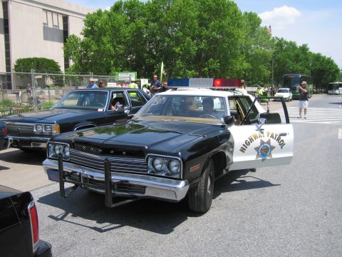1991 Ford LTD Crown Victoria - black-and-white Dodge Highway Patrol sedan with its door open sits among other vintage patrol cars on a street near trees.