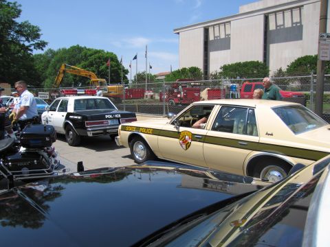 1991 Ford LTD Crown Victoria - tan-and-brown State Police sedan sits among other parked patrol cars and fire equipment in a lot beside a large building.
