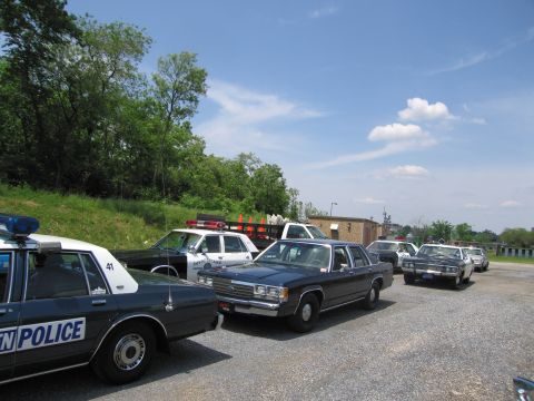 1991 Ford LTD Crown Victoria - row of vintage police cars, including one marked POLICE 41, sits parked on a gravel lot beside a grassy hill under a blue sky.