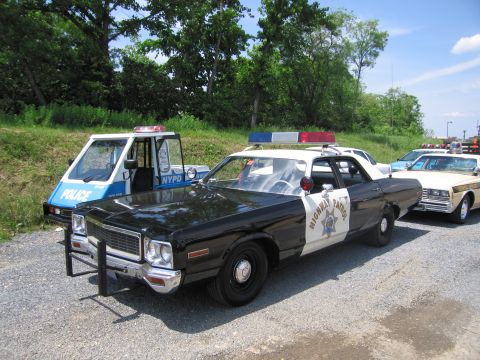 1991 Ford LTD Crown Victoria - black-and-white Dodge Highway Patrol sedan with a push bar is parked beside a blue-and-white NYPD car on a gravel lot.
