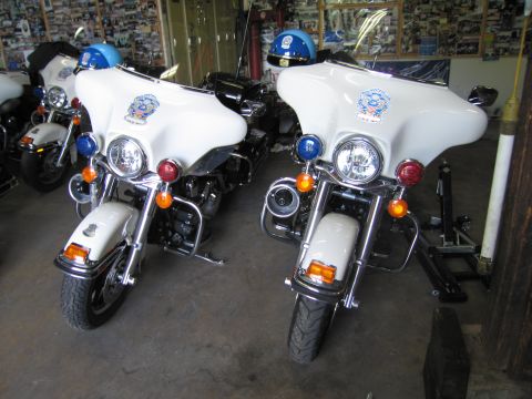 1991 Ford LTD Crown Victoria - Two white police touring motorcycles topped with blue helmets are parked front-on inside a garage hung with photos.