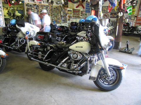 1991 Ford LTD Crown Victoria - black-and-white police touring motorcycle is parked among other bikes in a garage crowded with signs and memorabilia.