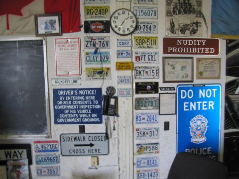 1991 Ford LTD Crown Victoria - wall is densely covered with vintage license plates, a clock, and signs reading DO NOT ENTER and NUDITY PROHIBITED.