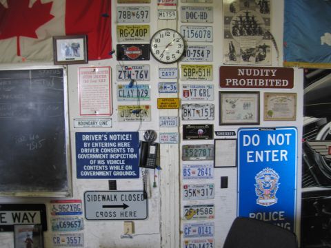 1991 Ford LTD Crown Victoria - cluttered wall covered in license plates and warning signs, with a Canadian flag at upper left and a DO NOT ENTER sign at right.
