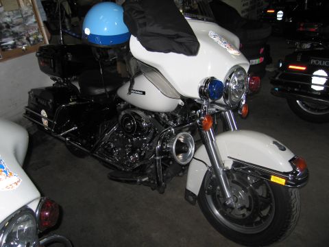 1991 Ford LTD Crown Victoria - white-and-black police touring motorcycle with a blue helmet on the seat, photographed front-left among other bikes in a garage.