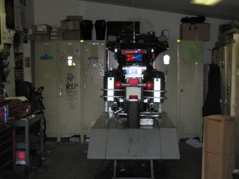 1991 Ford LTD Crown Victoria - Direct rear view of a police touring motorcycle with red emergency lights illuminated, raised on a lift in a dim garage with lockers.