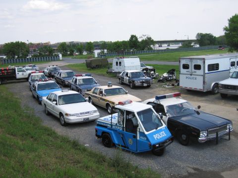 1991 Ford LTD Crown Victoria - Elevated view of a lot packed with assorted police cars, vans and trailers, with a blue NYPD three-wheeled scooter in the foreground.