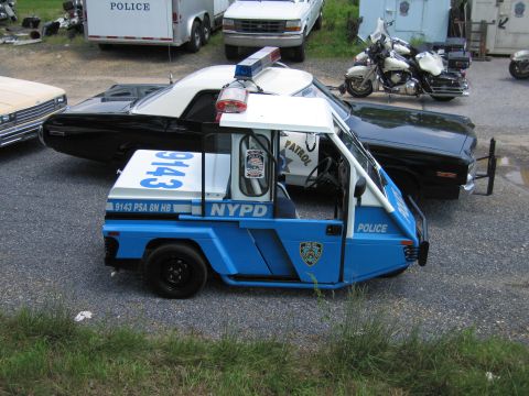 1991 Ford LTD Crown Victoria - blue NYPD three-wheeled police scooter parked beside a black muscle car and a police touring motorcycle on a gravel lot.