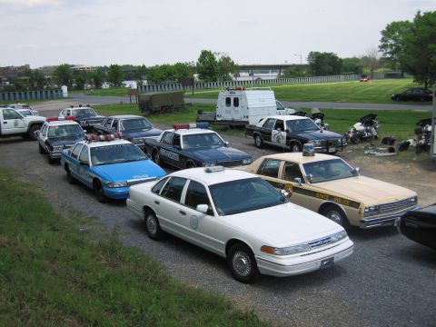1991 Ford LTD Crown Victoria - Elevated view of many parked police cars including a tan STATE POLICE sedan and blue cruisers, with motorcycles and trailers behind.