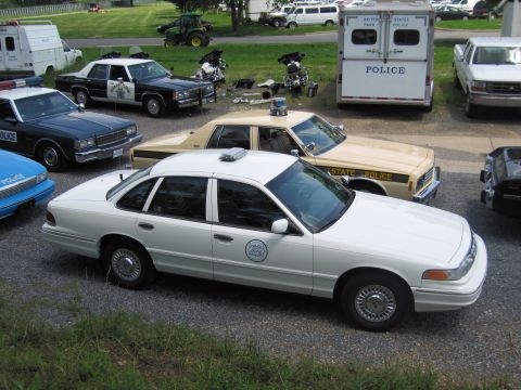 1991 Ford LTD Crown Victoria - Elevated view of a white police sedan in the foreground and a tan STATE POLICE sedan behind, among many cruisers on a gravel lot.