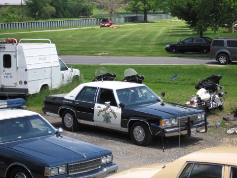1991 Ford LTD Crown Victoria - black-and-white HIGHWAY PATROL sedan parked on grass beside a police touring motorcycle, with a Park Police van to the left.