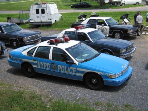 1991 Ford LTD Crown Victoria - blue-and-white NYPD police sedan marked POLICE parked on a gravel lot among other cruisers, vans and a Park Police trailer.