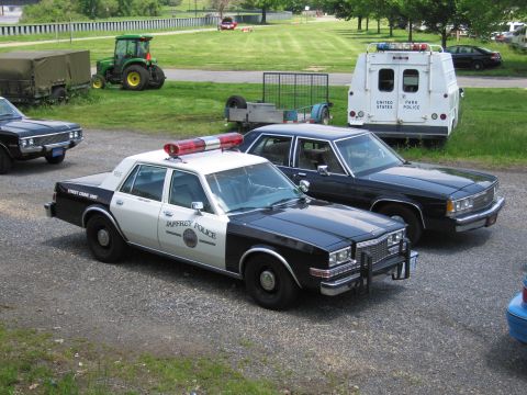 1991 Ford LTD Crown Victoria - black-and-white Jaffrey Police sedan and a black sedan park on gravel, with a green tractor and a United States Park Police truck in the ba…