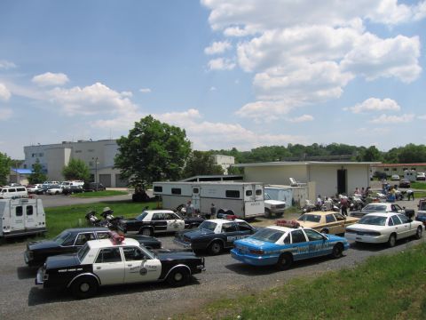 1991 Ford LTD Crown Victoria - wide view of a police-vehicle show with multiple cruisers, a blue NYPD car, trailers and motorcycles parked on gravel under a partly cloudy…