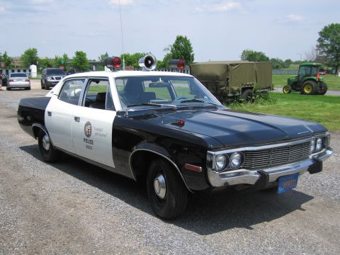 1991 Ford LTD Crown Victoria - black-and-white police sedan with roof lights, twin spotlights and a loudspeaker sits on gravel near a green military truck and a tractor.