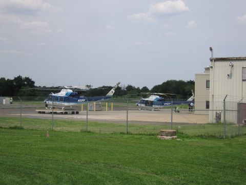 1991 Ford LTD Crown Victoria - Two blue-and-white helicopters sit on a fenced helipad next to a tan building, viewed across a grassy field under a hazy sky.