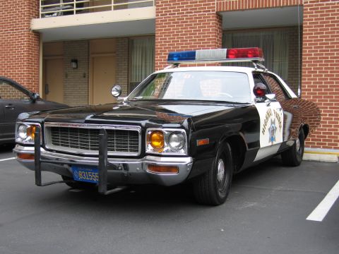 1991 Ford LTD Crown Victoria - black-and-white California Highway Patrol Dodge sedan with twin spotlights and a roof lightbar parks in front of a brick motel.
