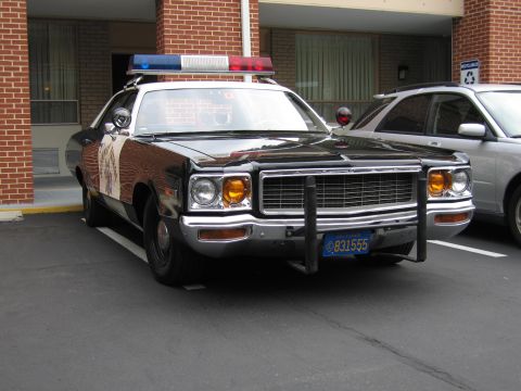 1991 Ford LTD Crown Victoria - black-and-white police Dodge sedan with twin spotlights, roof lights and a chrome push bar parks at a brick motel beside a silver car.