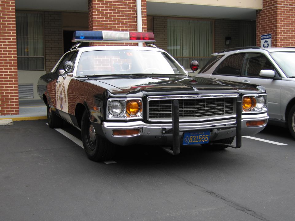 Front three-quarter of a black-and-white police Dodge sedan with twin spotlights and push bar parked at a brick motel