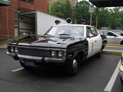 1991 Ford LTD Crown Victoria - black-and-white AMC Matador police sedan with twin spotlights and roof lights parks beside a brick building and a white box truck.