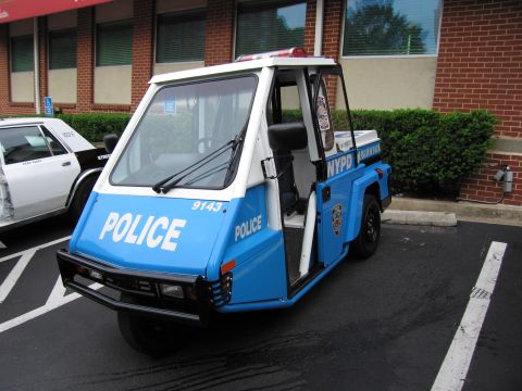 1991 Ford LTD Crown Victoria - blue-and-white NYPD three-wheeled police scooter marked POLICE 9143 sits parked in a striped lot beside a brick building.