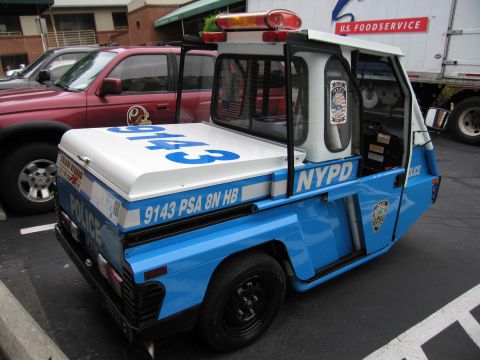1991 Ford LTD Crown Victoria - blue and white NYPD three-wheeled police scooter marked 9143 parked beside a red pickup and a U.S. Foodservice box truck.