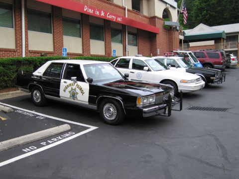 1991 Ford LTD Crown Victoria - black and white Highway Patrol Ford leads a row of marked police cars parked in front of a brick restaurant.