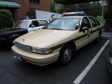 1991 Ford LTD Crown Victoria - tan Chevrolet Caprice marked State Police with a roof lightbar parked near a Guest Registration entrance.