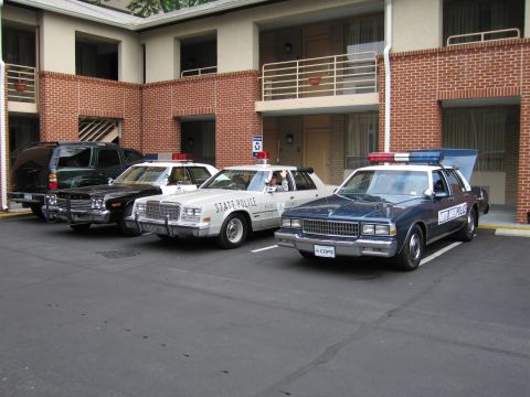 1991 Ford LTD Crown Victoria - Three vintage police cars, including a white State Police sedan and a dark sedan, parked in a row at a motel.
