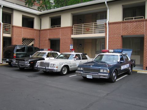 1991 Ford LTD Crown Victoria - row of classic police cars including a white STATE POLICE sedan and a dark sedan parked outside a brick motel.