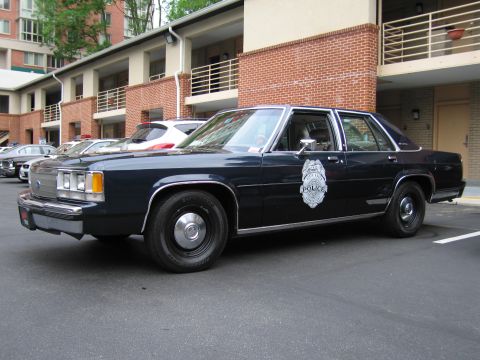 1991 Ford LTD Crown Victoria - dark blue Ford LTD Crown Victoria police sedan with a door badge parked along a brick motel walkway.