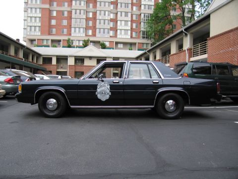 1991 Ford LTD Crown Victoria - Dark blue Ford LTD Crown Victoria police sedan shown in profile, parked before brick apartment buildings and a high-rise.