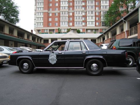 1991 Ford LTD Crown Victoria - Dark blue Ford LTD Crown Victoria police cruiser with a police door badge, parked among vehicles near a tall apartment building.