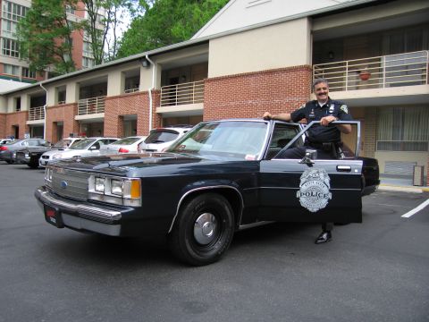1991 Ford LTD Crown Victoria - uniformed police officer poses at the open door of a dark blue Ford LTD Crown Victoria cruiser in a parking lot.