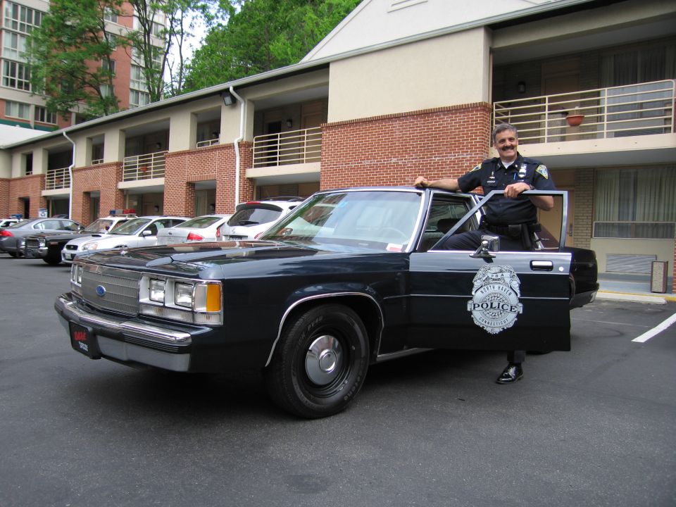 Uniformed officer stands by open door of dark blue Ford LTD Crown Victoria police car