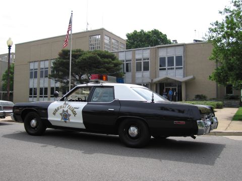 1991 Ford LTD Crown Victoria - black-and-white 1970s Highway Patrol sedan with roof light bar parked at a curb before a mid-century office building.