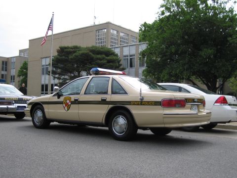 1991 Ford LTD Crown Victoria - Rear three-quarter view of a tan-and-brown Chevrolet Caprice State Police sedan parked near a flag and office building.