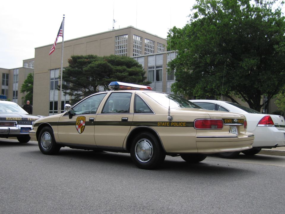 Rear three-quarter of tan Chevrolet Caprice State Police sedan by an office building