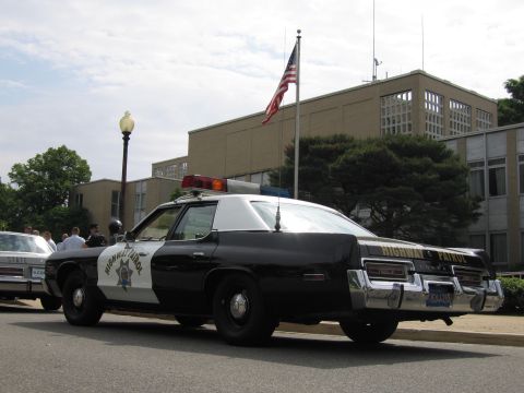 1991 Ford LTD Crown Victoria - Rear view of a black-and-white 1970s Highway Patrol sedan with roof light bar parked at a curb near a U.S. flag.