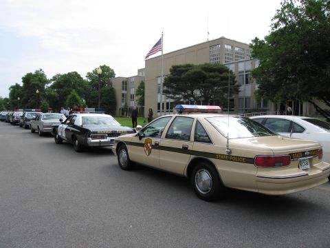 1991 Ford LTD Crown Victoria - lineup of police cars including a tan State Police Chevrolet Caprice in front of a mid-century office building.