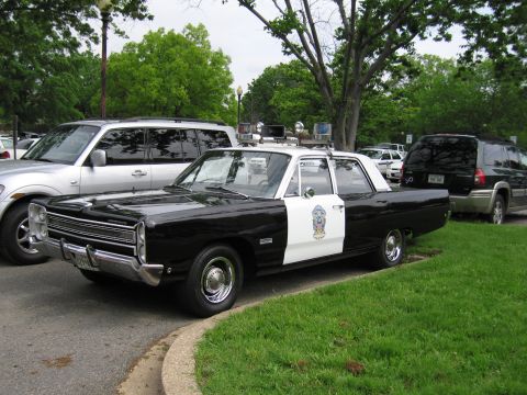 1991 Ford LTD Crown Victoria - black-and-white 1960s Plymouth police sedan with roof lights and a badge decal parked along a grassy lot edge.