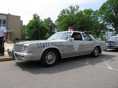 1991 Ford LTD Crown Victoria - silver Rhode Island State Police sedan with a front push bumper and roof lights parked beside a building, officer at left.
