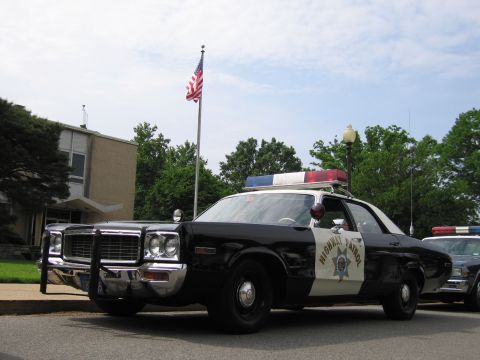 1991 Ford LTD Crown Victoria - Front view of a black-and-white 1970s Highway Patrol sedan with push bumper and roof lights parked near a U.S. flag.