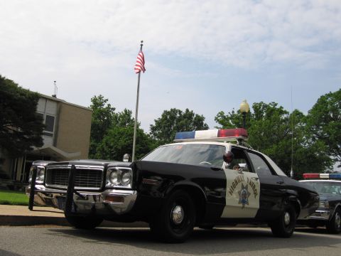 1991 Ford LTD Crown Victoria - Front view of a black-and-white 1970s Highway Patrol sedan with grille guard and roof lights parked near a U.S. flag.